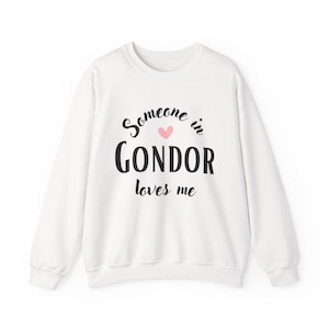 May include: White sweatshirt with the text "Someone in Gondor loves me" in black script. A small pink heart is above the word "Gondor". The sweatshirt has a crew neck and long sleeves.