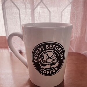 May include: White ceramic mug with a curved handle. The mug features a black and white graphic of a grumpy character inside a circular frame with the text "GRUMPY BEFORE MY COFFEE". The mug is on a wooden surface.
