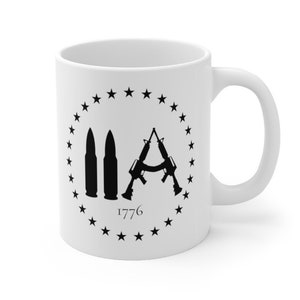 May include: White ceramic coffee mug featuring a black silhouette of two rifles crossed and two bullets, encircled by a ring of stars. The text "1776" is positioned beneath the image.
