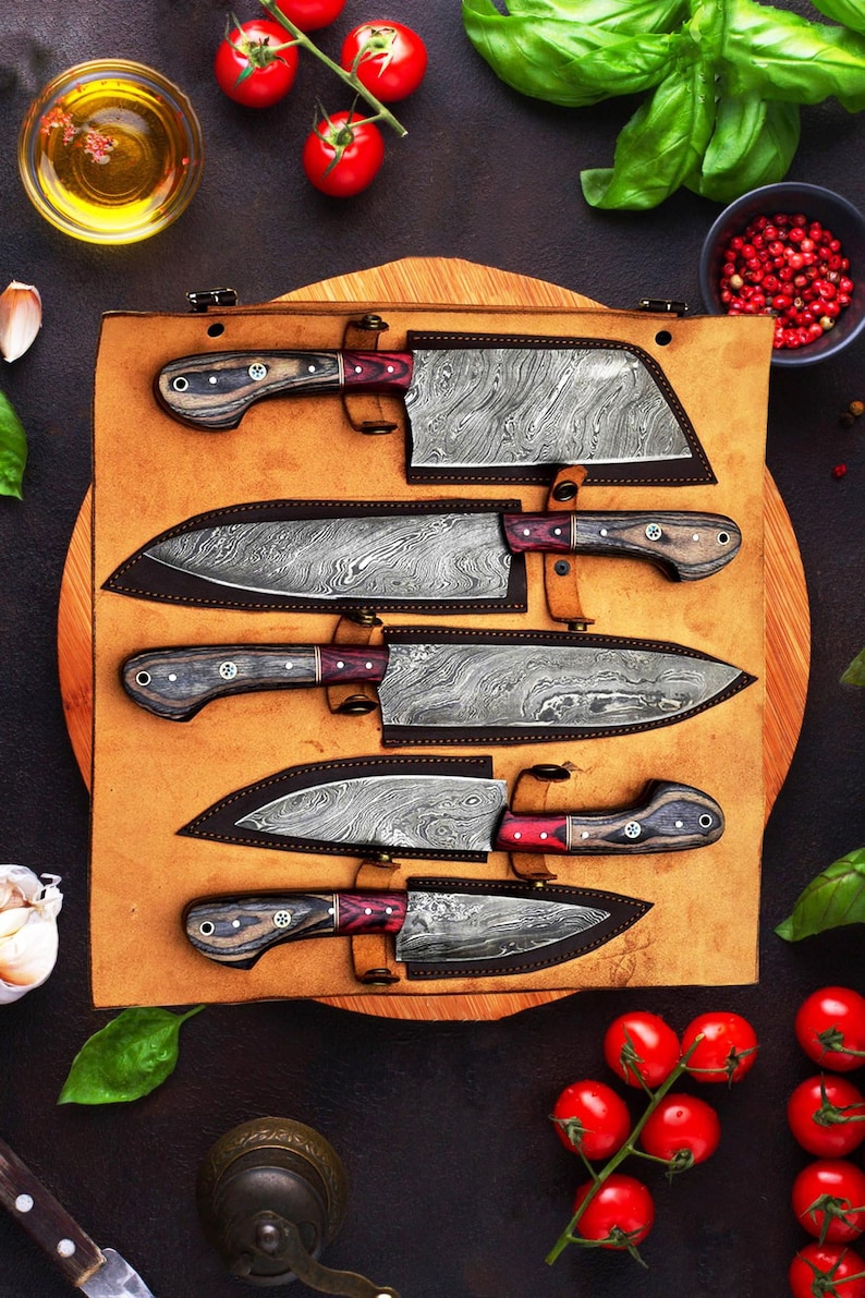 May include: A set of five kitchen knives with Damascus steel blades and dark wood handles with red accents, each stored in a custom-fitted brown leather roll. The knives feature distinct wavy patterns on the metal blades and are arranged horizontally.