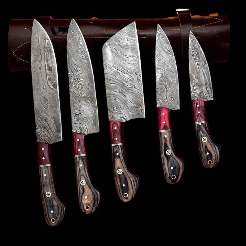 May include: A set of five kitchen knives with Damascus steel blades featuring a wavy pattern. Each knife has a handle made of dark wood with red accents, secured by three metal rivets. The knives are arranged in front of a brown leather roll.