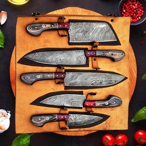 May include: A set of five kitchen knives with Damascus steel blades and dark wood handles with red accents, each stored in a custom-fitted brown leather roll. The knives feature distinct wavy patterns on the metal blades and are arranged horizontally.