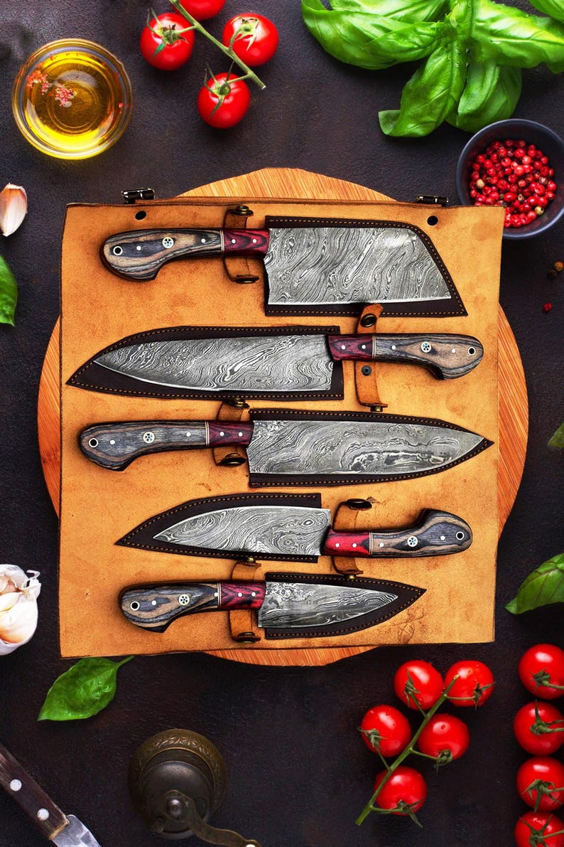 May include: A set of five kitchen knives with Damascus steel blades and dark wood handles with red accents, each stored in a custom-fitted brown leather roll. The knives feature distinct wavy patterns on the metal blades and are arranged horizontally.