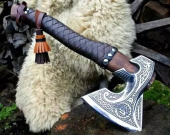 May include: A handcrafted axe with a wooden handle and a silver-colored, engraved blade. The handle is wrapped in dark brown leather with a braided pattern. A leather tassel hangs from the end of the handle. The axe is displayed on a wooden surface.