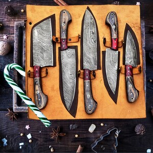 May include: A set of five Damascus steel knives with patterned blades and wooden handles, each secured in a tan leather roll. The knives feature distinct blade shapes, including a cleaver and various chef knives, arranged on a flat surface.
