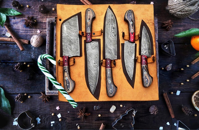 May include: A set of five Damascus steel knives with patterned blades and wooden handles, each secured in a tan leather roll. The knives feature distinct blade shapes, including a cleaver and various chef knives, arranged on a flat surface.