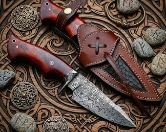 May include: A fixed-blade knife with a dark reddish-brown wooden handle and a patterned Damascus steel blade. The knife is accompanied by a brown leather sheath featuring a cross-stitched detail and a textured black inlay, resting on a carved wood surface.