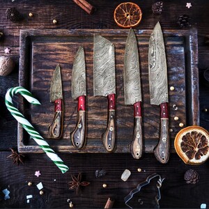 May include: A set of five knives with patterned Damascus steel blades and wooden handles featuring red accents. The knives are arranged in a row on a dark wooden tray, accompanied by a green and white candy cane and dried orange slices.