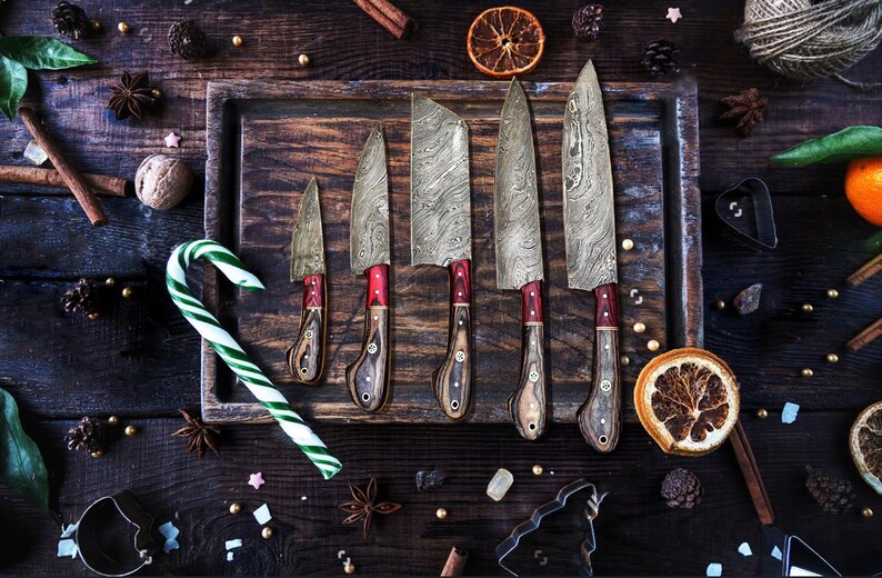 May include: A set of five knives with patterned Damascus steel blades and wooden handles featuring red accents. The knives are arranged in a row on a dark wooden tray, accompanied by a green and white candy cane and dried orange slices.