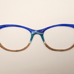 May include: Blue cat-eye glasses with a beach scene painted on the frames. The scene includes blue water, white waves, and brown sand. There are gold anchor and star charms on the temples.