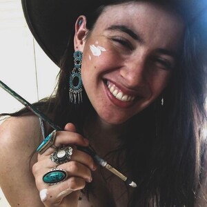Può includere: Una donna indossa un cappello a tesa larga, orecchini turchesi e diversi anelli con pietre turchesi. Sta tenendo un pennello e sorridendo.