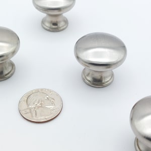 May include: Brushed nickel cabinet knobs with a quarter for size comparison. The knobs have a rounded top and a decorative base. The quarter shows the profile of George Washington and the words "Liberty" and "2022".