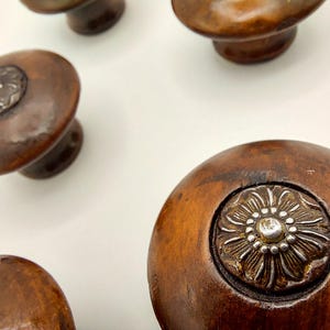 May include: Close-up of several antique wooden cabinet knobs. The knobs are a warm brown color with a silver floral design in the center. The knobs have a rounded shape and are likely for furniture.