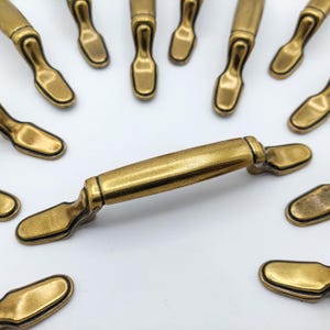 May include: A collection of antique brass cabinet pulls. The pulls have a rectangular bar with rounded ends and a curved handle. The hardware is a warm, golden color, suggesting an aged finish. The pulls are arranged on a white background.