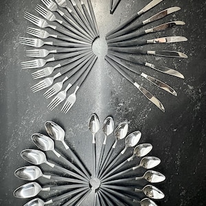 May include: A flatware set featuring stainless steel heads and matte black handles arranged in circular patterns. The collection includes forks, knives, and spoons, all displaying a consistent design with metallic silver tops and dark, slender handles.