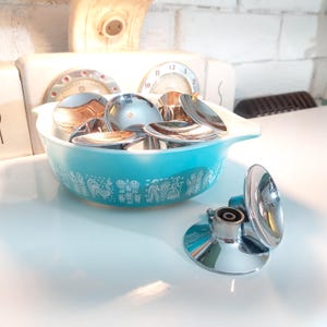 May include: A collection of shiny, silver spoons in a turquoise Pyrex bowl with a white decorative pattern. A chrome-plated, round-faced object sits nearby. The background includes a white brick wall and a vintage kitchen appliance.