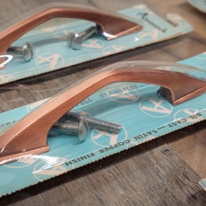 May include: Two copper cabinet pulls with a satin finish. The pulls are 1 inch in length and are packaged on a blue and white card with the text "Die Cast Copper Satin Finish" and "Pull 1" inch Acrylic".