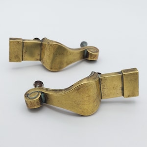 May include: Two antique brass furniture feet with a rectangular base and a curved, ornate design. Each foot has a decorative scroll detail and a screw for attachment. The brass has a warm, aged patina.