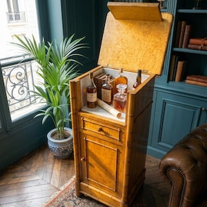 May include: A tall, light brown wooden bar cabinet with a faux bamboo frame, featuring a hinged top lid that opens to reveal a white marble surface. The cabinet includes a single drawer and a lower cupboard door, holding several glass liquor bottles.