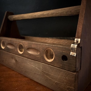 May include: A dark stained wooden toolbox with a handle and a series of circular and oval cutouts. The toolbox is constructed with visible wood grain and metal hardware. The design suggests a rustic or vintage aesthetic.