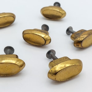 May include: Several antique brass oval cabinet knobs with silver screws are scattered on a white surface. The knobs have a worn, aged appearance, with a slightly tarnished gold finish. These vintage hardware pieces are ideal for furniture restoration.
