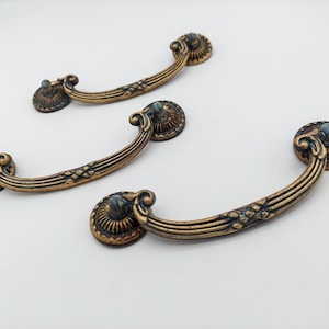 May include: Three curved drawer pulls with an antique brass finish. Each handle features a central ribbed design with cross-hatch detailing and circular mounting plates at both ends, decorated with radial grooves and a dark, weathered patina.