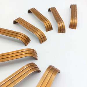 May include: A collection of vintage bronze-toned cabinet pulls. Each pull has a curved design with three horizontal grooves. The hardware is arranged on a white surface, showcasing their aged appearance and potential for furniture restoration projects.