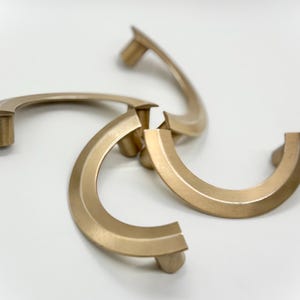 May include: Four gold-colored cabinet pulls arranged on a white surface. Each pull has a curved, semi-circular design with a rectangular base. The pulls are made of metal and have a matte finish, suitable for furniture hardware.
