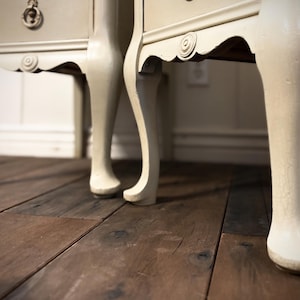 May include: Close-up of two antique cream-colored bedside tables with curved legs. The tables have decorative scrollwork and a distressed finish. The wooden floorboards are a rich, dark brown, creating a contrast with the light-colored furniture.