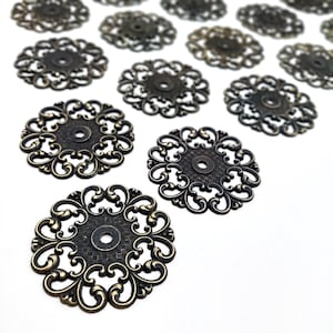 May include: A collection of circular metal filigree findings in an antique bronze finish. Each piece features a central hole surrounded by a textured grid pattern and an intricate, openwork scroll design along the outer edge of the circular shape.