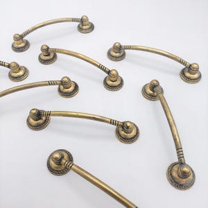 May include: A collection of antique brass drawer pulls. Each pull features a curved handle with decorative rope detailing and circular mounting plates. The hardware has a vintage aesthetic, suitable for furniture restoration or adding a classic touch to cabinetry.