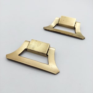 May include: Two brass-colored metal drawer pulls with a trapezoidal shape and a hinged drop handle. Each piece features a flat rectangular mounting plate at the top connected to a wider, flared base bar that swings freely from the central hinge point.