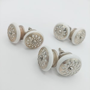 May include: A collection of white and silver cabinet knobs. Each knob features a white ceramic base with a silver floral design and a silver metal stem. The knobs are arranged on a white surface.