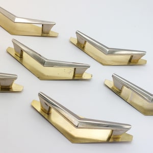 May include: Set of six mid-century modern cabinet pulls. Each pull features a gold-toned base with a silver-toned handle. The pulls have a distinctive V-shape design, adding a retro touch to furniture. Ideal for updating drawers or cabinets.