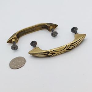 May include: Two antique brass drawer pulls with mounting screws. One pull has a leaf design, the other has the number "F2643" stamped on it. A US quarter is included for size comparison. The pulls are curved and have a vintage aesthetic.