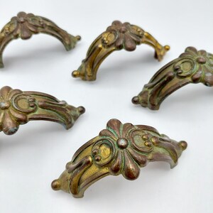 May include: A collection of antique brass drawer pulls with a floral design. The pulls have a curved shape and are finished in a distressed gold and green patina. The hardware is likely for furniture restoration or decorative use.