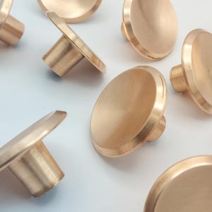 May include: A collection of round, bronze-colored cabinet knobs with a flat, disc-shaped face and a cylindrical stem. The knobs are arranged on a white surface, with some angled to show the stem.