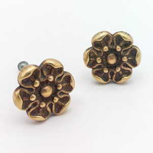 May include: Two antique brass flower-shaped cabinet knobs. Each knob features a six-petal design with a central circular detail. The knobs have a vintage, aged appearance, with a warm, golden-brown color. They are likely used for furniture or decorative purposes.