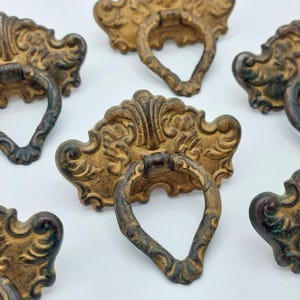 May include: Ornate, antique-style drawer pulls with a distressed gold finish. Each pull features a decorative backplate with scrolling details and a ring handle. The hardware is likely made of metal and adds a vintage touch to furniture.