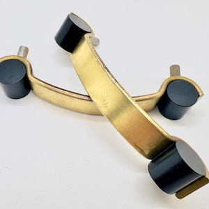 May include: Two curved drawer pulls featuring a brushed gold metal finish and cylindrical black end caps. Each handle has two silver-toned mounting posts extending from the back, designed for attachment to furniture surfaces or cabinet doors.