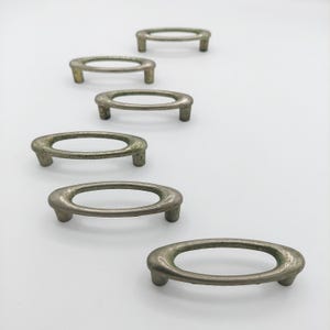 May include: A set of six oval-shaped metal drawer pulls arranged in a diagonal line. Each pull features a smooth, rounded frame with a weathered, muted silver-toned finish and two short, cylindrical mounting posts extending from the underside.