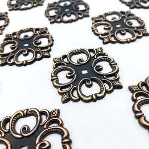 May include: A collection of square metal embellishments featuring an openwork scroll design. Each piece has a matte black center with raised, antiqued bronze-colored edges and a small square hole in the middle of the decorative pattern.