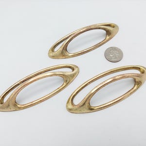 May include: Three oval-shaped brass drawer pulls with an open center design. Each handle features a smooth, metallic finish with subtle signs of wear. A United States quarter coin is placed near the handles to provide a sense of scale.