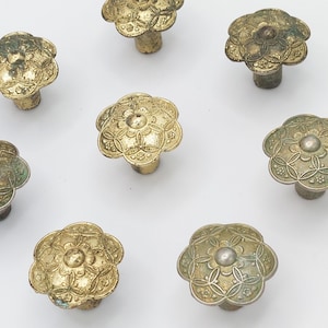 May include: A collection of antique brass flower-shaped cabinet knobs. Each knob features a detailed floral design with a distressed, aged finish. The knobs are arranged on a white surface, showcasing their intricate patterns and vintage appeal.