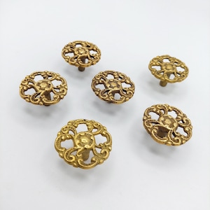 May include: A set of six round cabinet knobs in a metallic gold finish. Each knob features an intricate, openwork filigree design with a central circular element and swirling scrollwork patterns radiating outward to the outer edge of the knob.