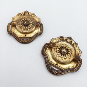 May include: Two gold-toned metal drawer pulls with a circular central medallion featuring a floral petal design. Each pull has a dark, weathered, and textured drop handle attached to an octagonal backplate with decorative scrollwork at the top.