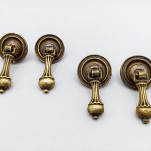 May include: Four antique brass-toned metal drawer pulls, each featuring a circular backplate and a hinged, teardrop-shaped drop handle. The hardware has a textured, weathered finish with visible dark patina in the recessed areas of the metal.