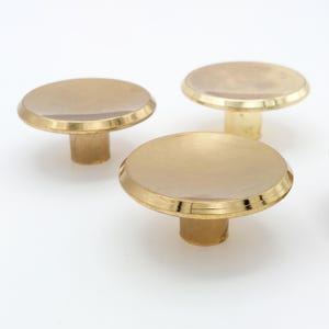 May include: Three brass cabinet knobs with a round, flat top and a short, cylindrical stem. The knobs have a polished, reflective surface and a warm, golden color. They are set against a plain white background.