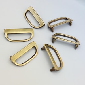 May include: Several brass-toned D-ring handles are arranged on a white surface. The handles have a curved, rectangular shape with a central opening. Some handles have mounting posts.
