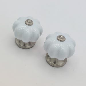 May include: Two white ceramic cabinet knobs with a pumpkin-like design. Each knob has a silver-colored base and a decorative silver center. These knobs are ideal for adding a touch of elegance to furniture.